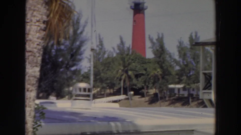 1966 Puerto Rico 8mm frame showing the red lighthouse, tropical trees, a road, and a small pale vehicle or trailer near the center.