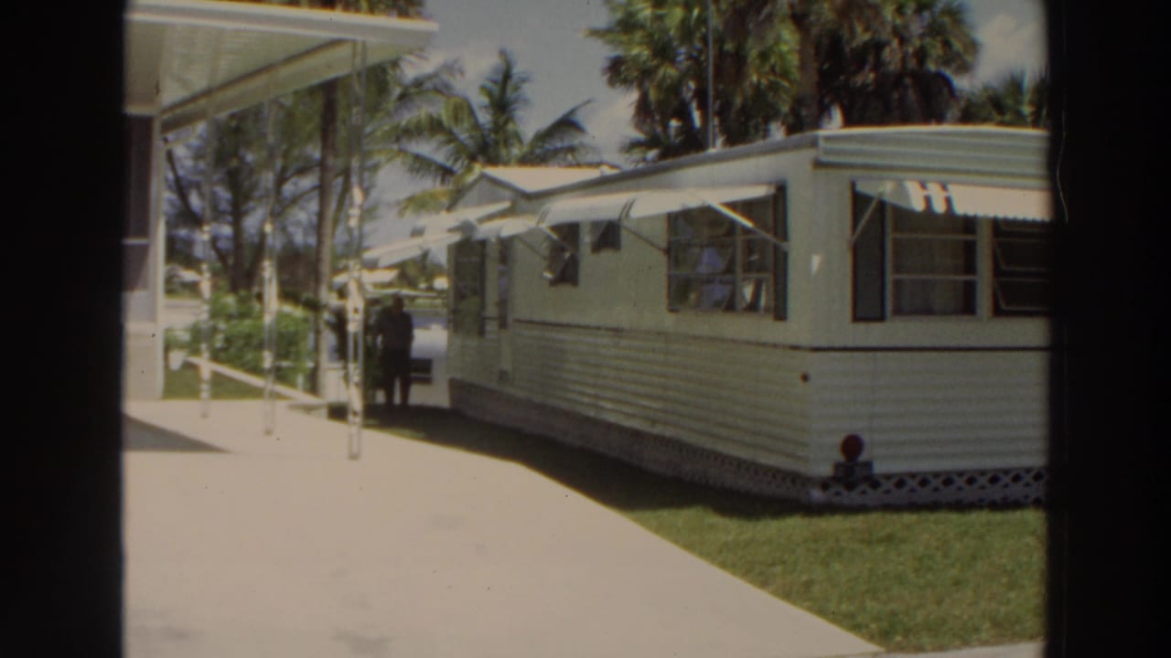 Elderly man walking near a white mobile home with awnings and palm trees.