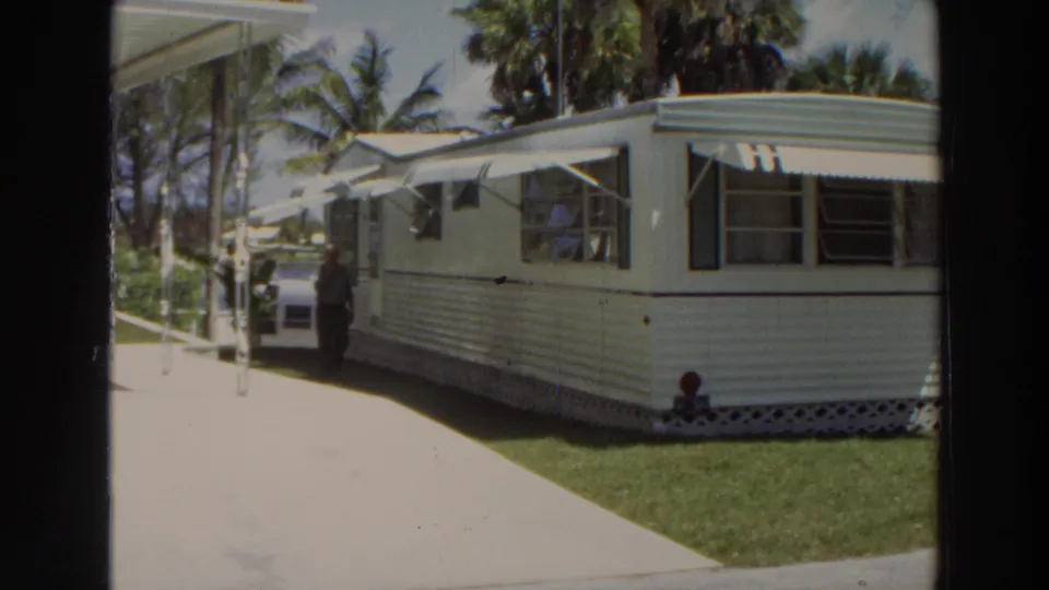 1966 Puerto Rico frame showing a man beside a white mobile home with striped siding, awnings, concrete path, lawn, and tropical trees.