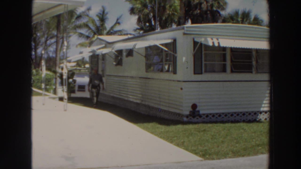 1966 Puerto Rico home-movie frame showing a white mobile home with green trim, awnings, a concrete drive, palm trees, and a parked car.