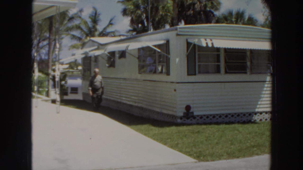 1966 Puerto Rico frame with an adult man walking beside a white and green mobile home, with awnings, palms, grass, and a car nearby.