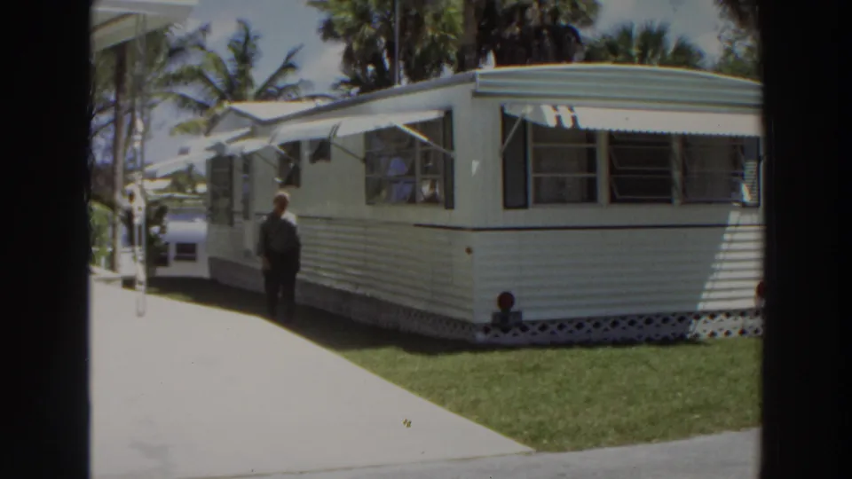 1966 Puerto Rico home-movie still showing a man in a light shirt and dark pants walking past a white mobile home with green trim.