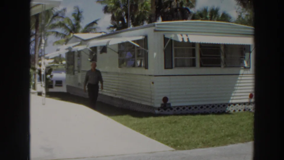1966 Puerto Rico frame of an adult man walking toward the camera area beside a white mobile home with green trim and striped awnings.