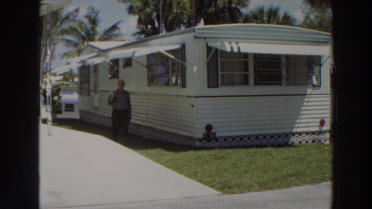 In 1966 Puerto Rico, a man walks near a white mobile home with metal awnings, striped siding, lawn, and palm trees.
