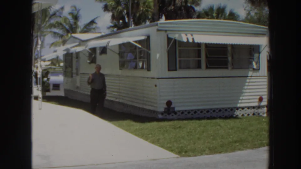 In 1966 Puerto Rico, a man walks near a white mobile home with metal awnings, striped siding, lawn, and palm trees.