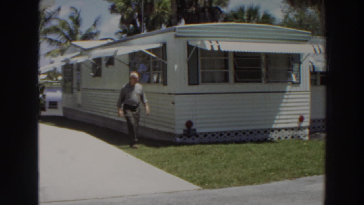 The man moves along the mobile home perimeter in Puerto Rico, with open awning windows, lawn, driveway, and palms visible.