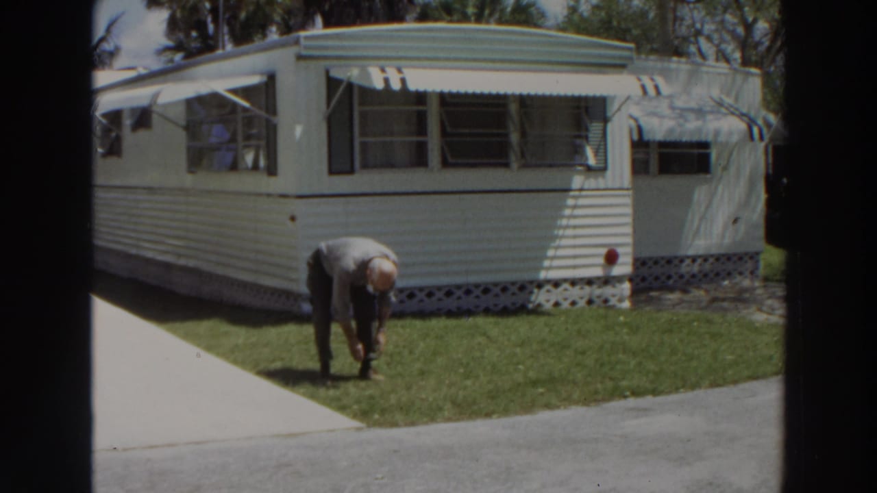 Man bending on a lawn beside a white mobile home and concrete walkway.