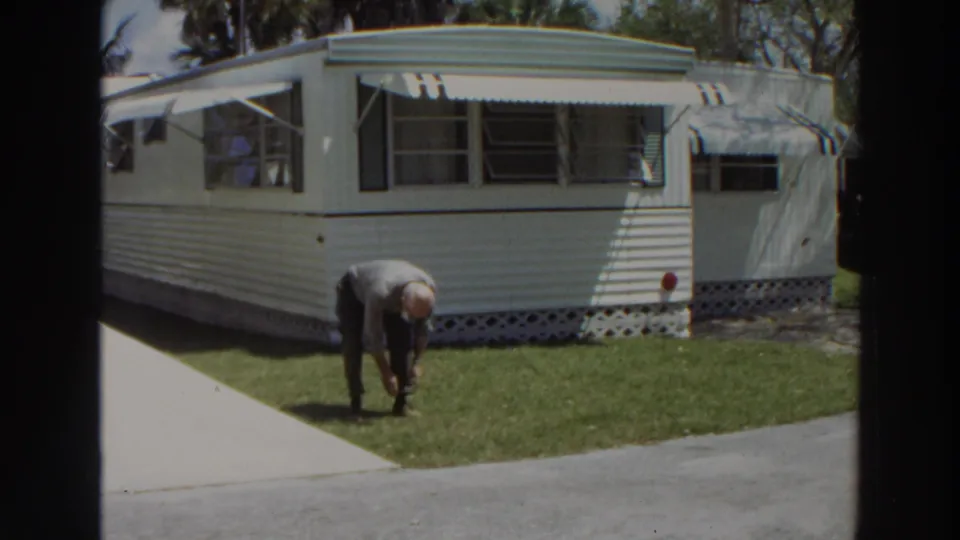 In 1966 Puerto Rico home-movie footage, a man bends near a lawn in front of a white mobile home with striped awnings.
