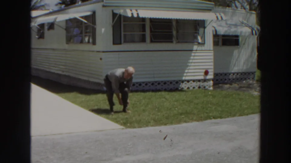 The man stands partly bent on the grass outside a white mobile home, with awnings and open windows behind him.
