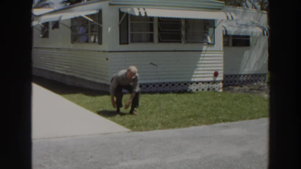 The man crouches lower near the edge of the grass, with the white mobile home and shaded windows in the background.