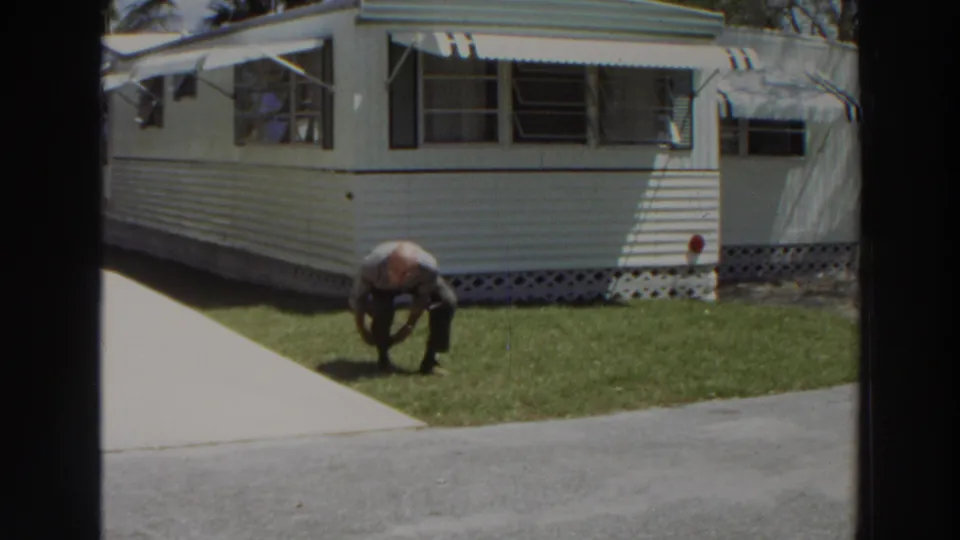 The man bends deeply toward his footwear on a sunny lawn outside a white mobile home in Puerto Rico home-movie footage.