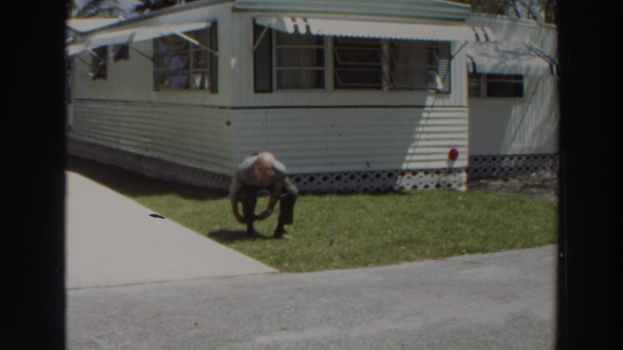 Man bends on a lawn near a white mobile home and concrete driveway.