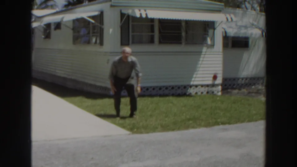 A man stands on the grass facing the camera outside a white mobile home in 1966 Puerto Rico home-movie footage.