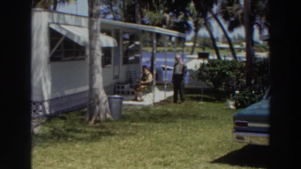 In a wide Puerto Rico home-movie frame, the man stands near a seated woman outside a white mobile home with palm trees and a partial car.