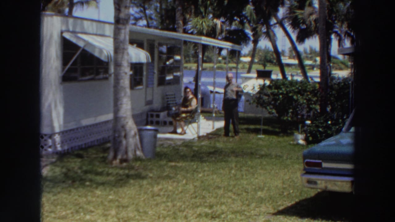 1966 Puerto Rico home-movie frame showing two adults relaxing outside a mobile home with palm trees, grass, shrubs, and a parked car edge visible.