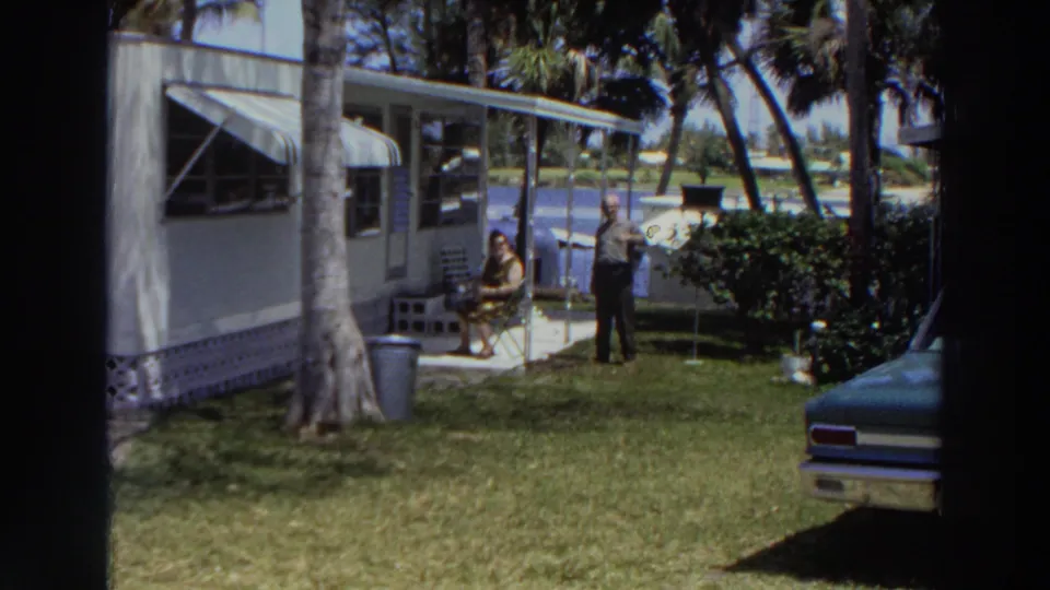 1966 Puerto Rico home-movie frame showing two adults relaxing outside a mobile home with palm trees, grass, shrubs, and a parked car edge visible.