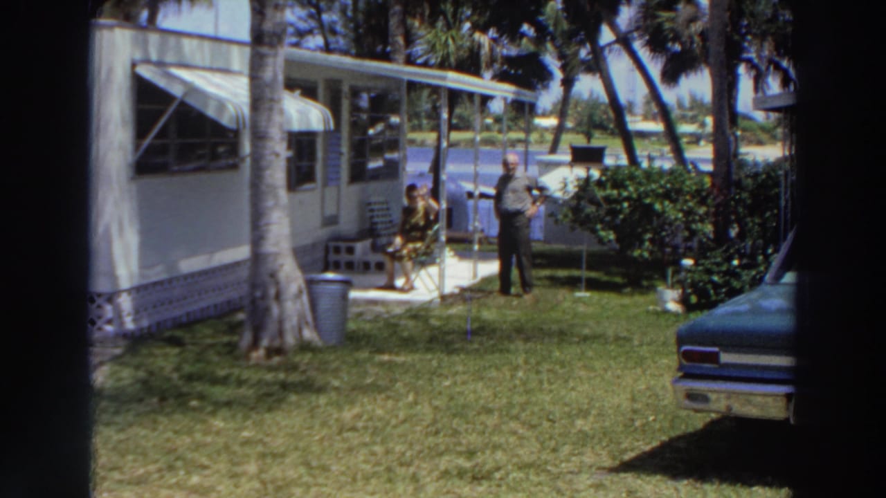 1966 Puerto Rico frame with a man standing and a woman seated near a mobile home, with awnings, palms, lawn, shrubs, and water beyond the yard.