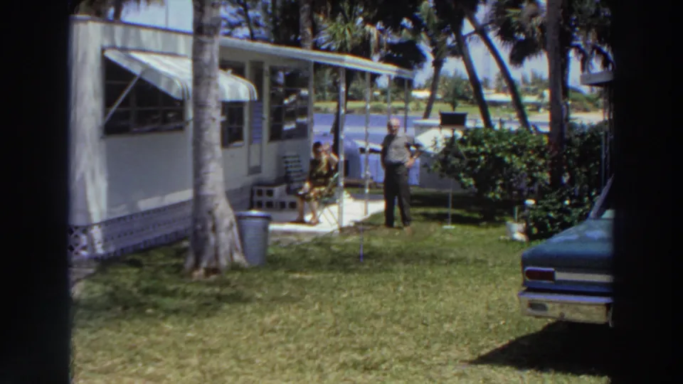 1966 Puerto Rico frame with a man standing and a woman seated near a mobile home, with awnings, palms, lawn, shrubs, and water beyond the yard.