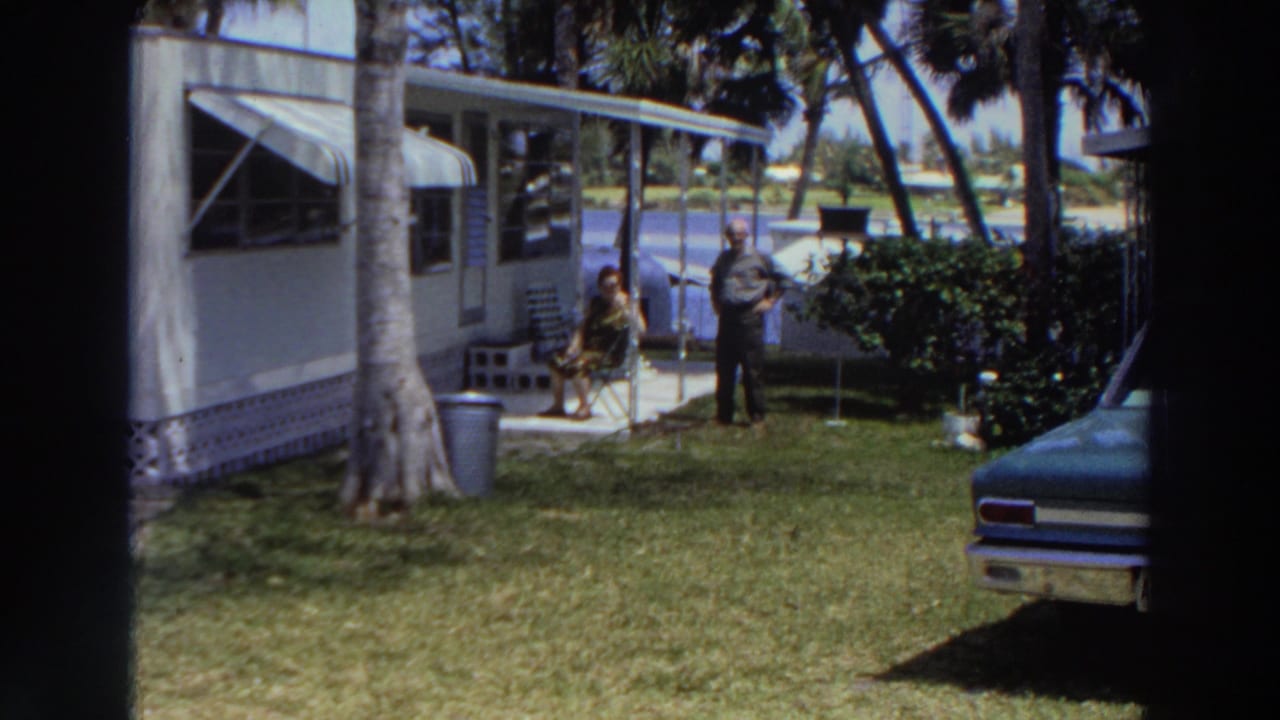 1966 Puerto Rico home-movie frame showing two adults in a grassy yard beside a mobile home, with palms, shrubs, water, and part of a vintage car.