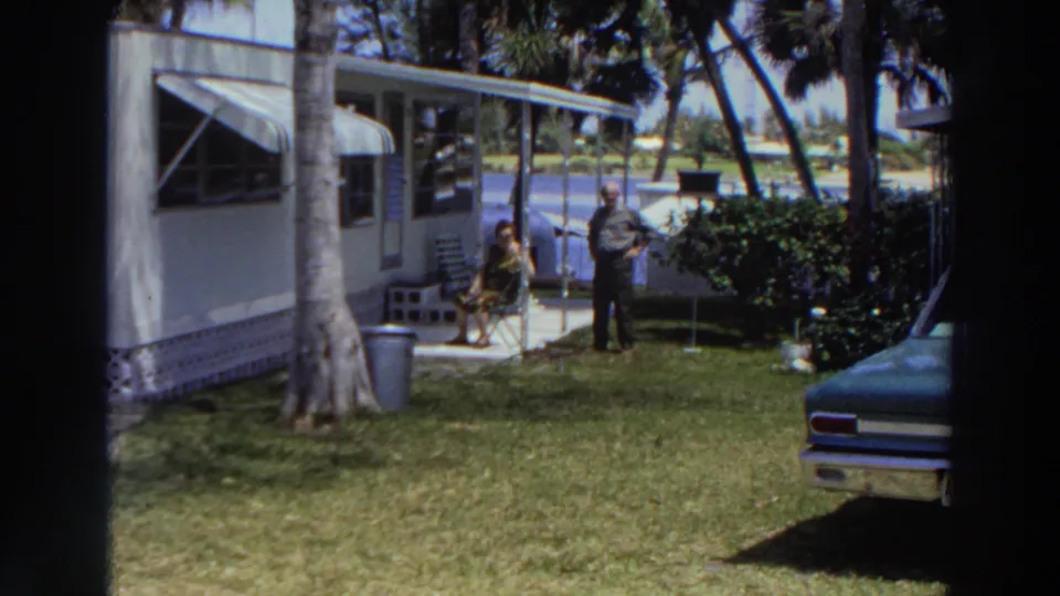 1966 Puerto Rico home-movie frame showing two adults in a grassy yard beside a mobile home, with palms, shrubs, water, and part of a vintage car.