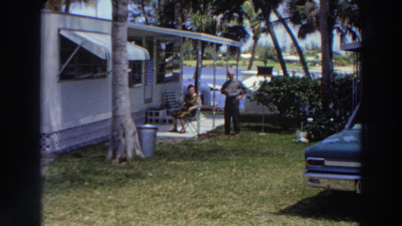 1966 Puerto Rico frame showing a man standing and a woman seated beside a mobile home, with a palm-lined lawn, shrubs, water, and car front nearby.