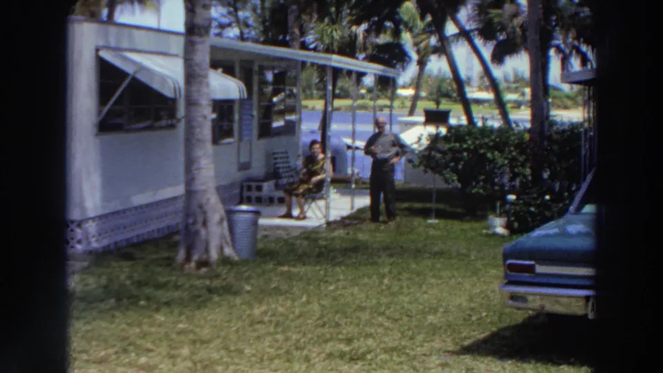 1966 Puerto Rico frame showing a man standing and a woman seated beside a mobile home, with a palm-lined lawn, shrubs, water, and car front nearby.