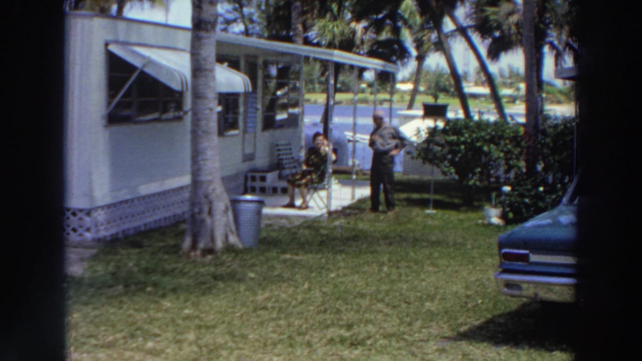 1966 Puerto Rico home-movie frame showing a woman seated outside a mobile home and a man standing nearby in a tropical yard.