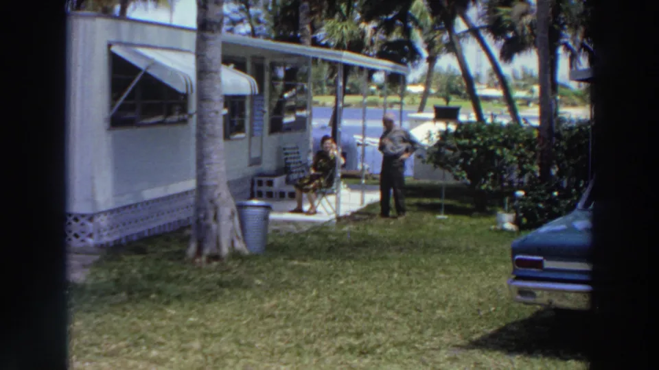 1966 Puerto Rico home-movie frame showing a woman seated outside a mobile home and a man standing nearby in a tropical yard.
