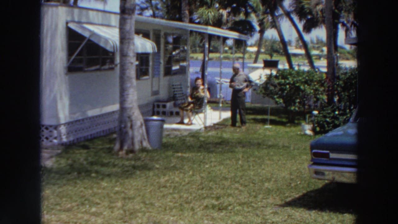 1966 Puerto Rico frame of a couple in a tropical yard beside a mobile home, with palm trees and part of a classic car visible.