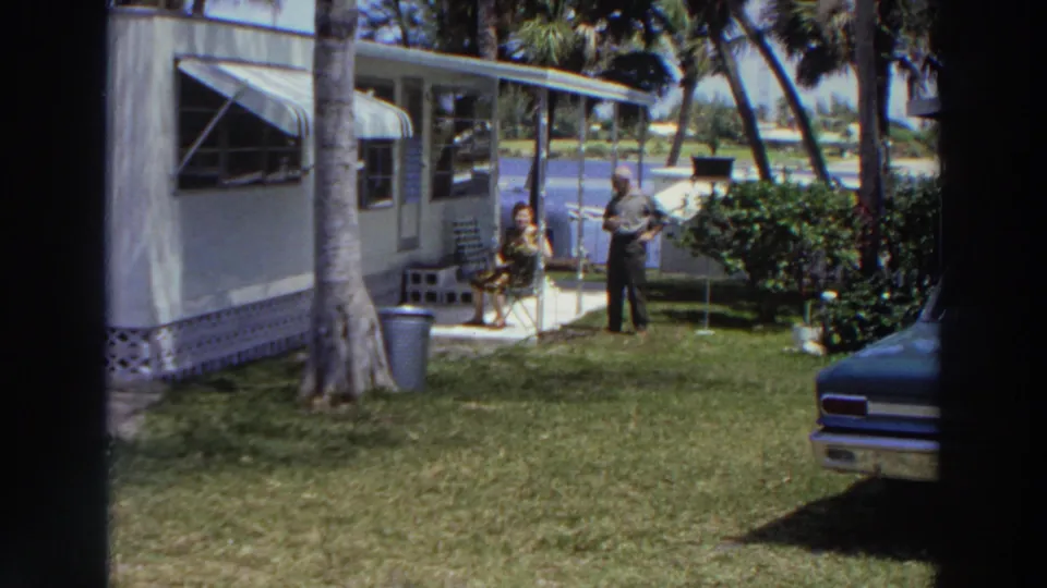1966 Puerto Rico frame of a couple in a tropical yard beside a mobile home, with palm trees and part of a classic car visible.