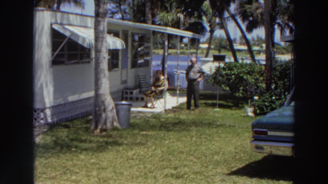 1966 Puerto Rico home-movie view of a tropical mobile-home yard with a seated woman, standing man, palms, lawn, and parked car.