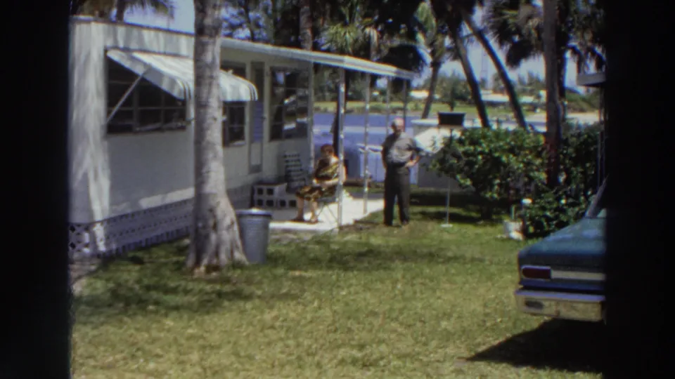 1966 Puerto Rico home-movie view of a tropical mobile-home yard with a seated woman, standing man, palms, lawn, and parked car.