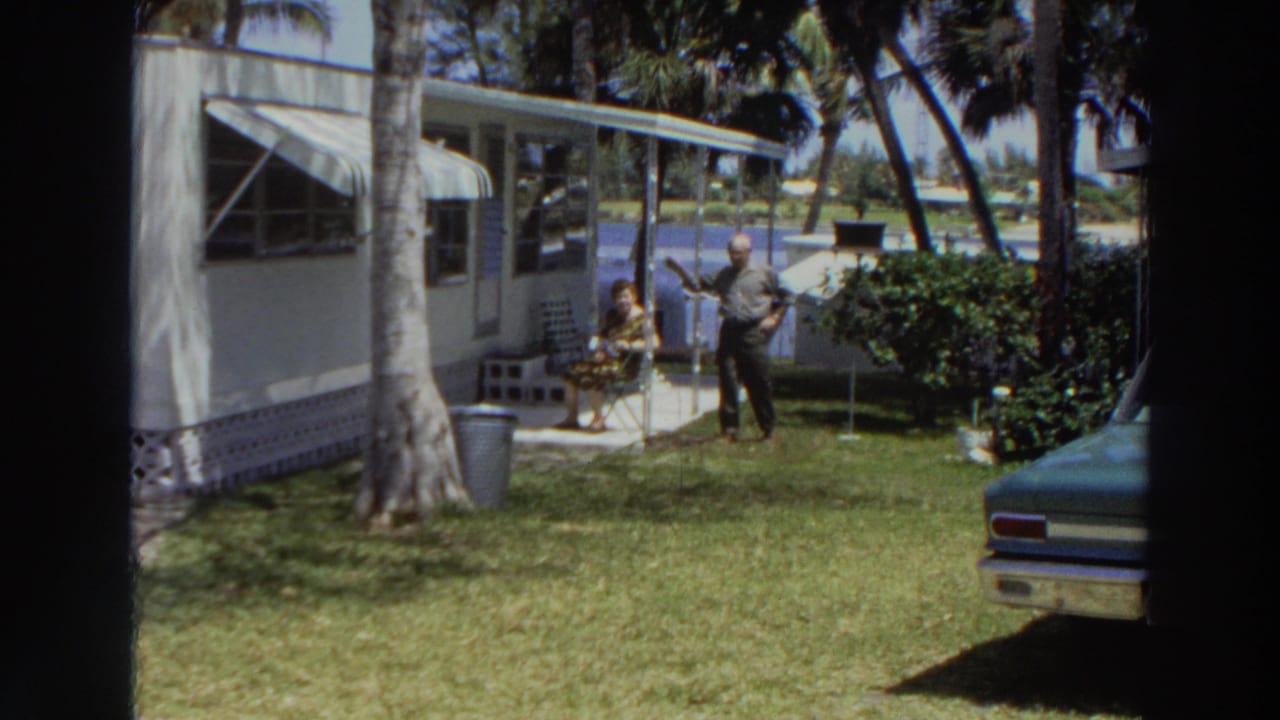 1966 Puerto Rico frame showing a man raising an arm near a seated woman outside a mobile home, with palms and a parked car in view.