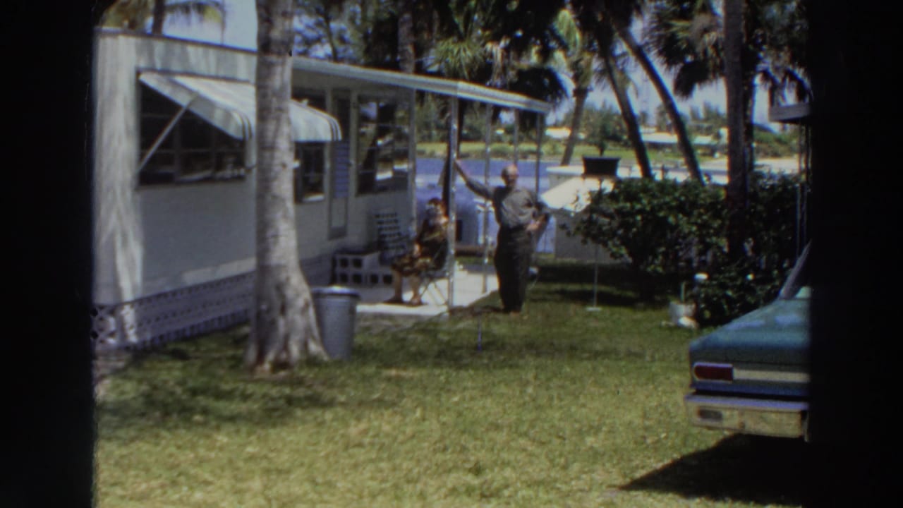 A white mobile home beside palm trees with a woman seated outside and a man standing on the lawn.