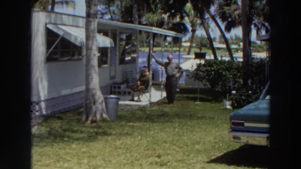 1966 Puerto Rico home-movie frame showing a white trailer, palm trees, a seated woman, and a man standing in the grassy yard.