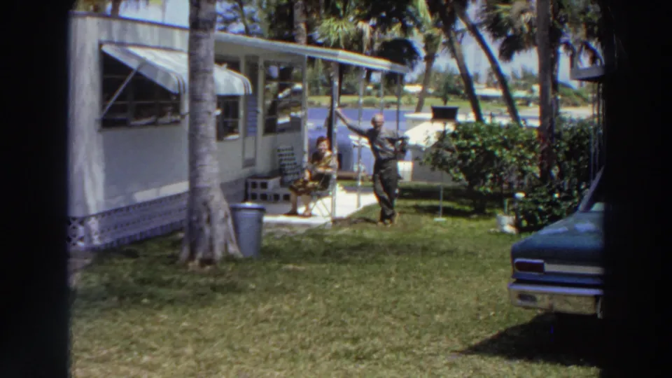 A 1966 Puerto Rico frame with a man in dark clothing standing outside a white mobile home as a woman sits nearby.