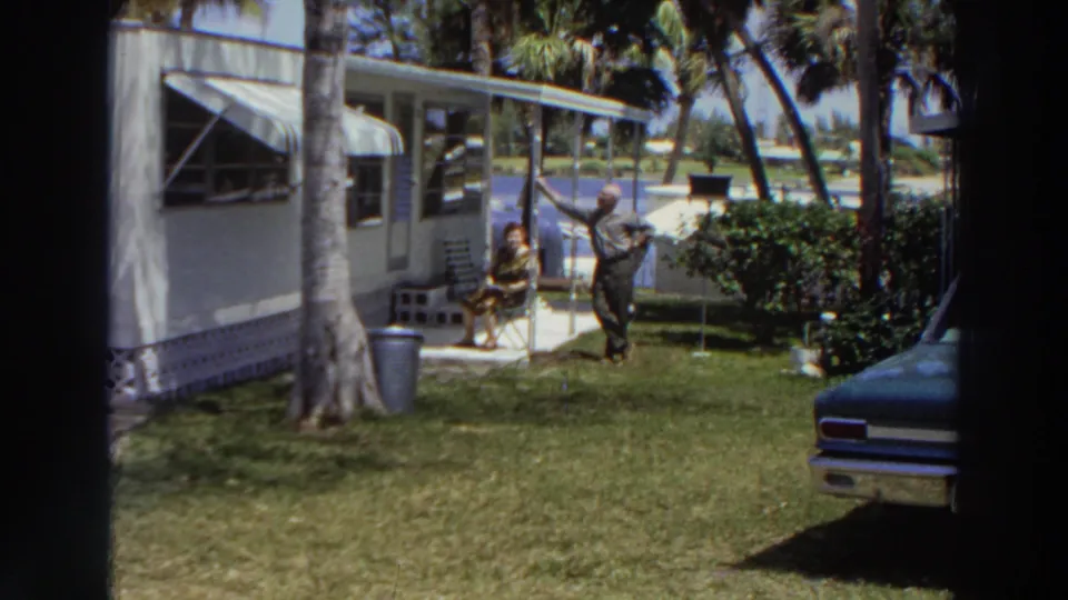 Wide 1966 Puerto Rico home-movie view of a mobile home yard with palms, a seated woman, a standing man, and a partial car.