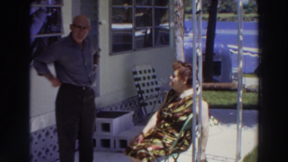 Closer 1966 Puerto Rico frame showing an adult man and seated woman outside a white mobile home, with patio furniture and lawn behind them.