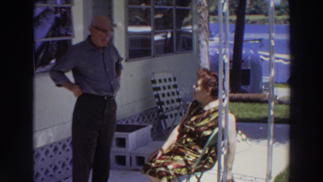 Man stands on a porch while a woman sits in a folding chair beside him.