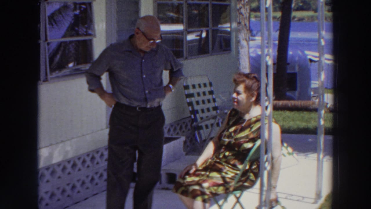 Older man standing beside seated older woman on a sunlit porch.