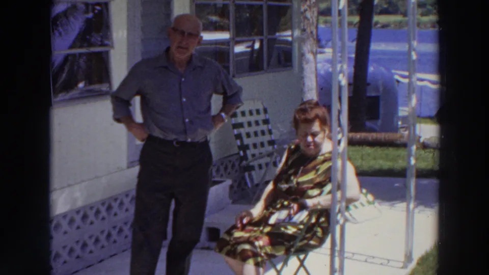 The man stands near the house as the seated woman looks down, possibly toward her lap or wrist, in this brief 1966 Puerto Rico porch scene.