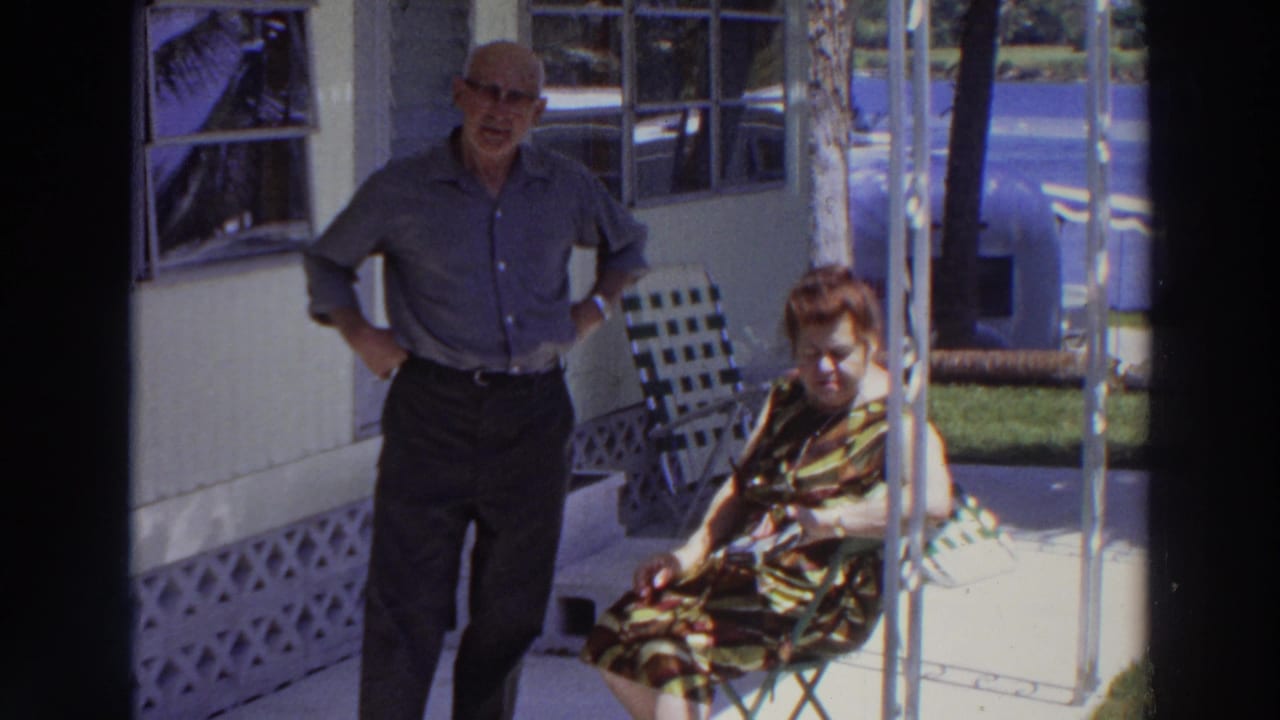 Older man standing beside seated woman on a shaded porch, viewed through a dark screen-door frame.