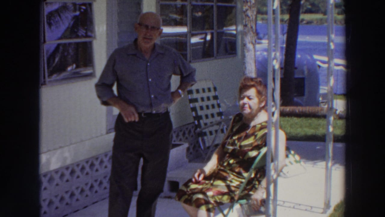 1966 Puerto Rico frame showing an older couple on a porch, with windows, lawn, and a partially visible blue vehicle beyond them.