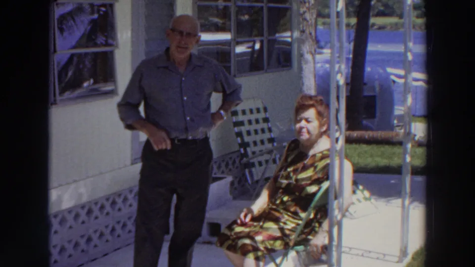 1966 Puerto Rico frame showing an older couple on a porch, with windows, lawn, and a partially visible blue vehicle beyond them.