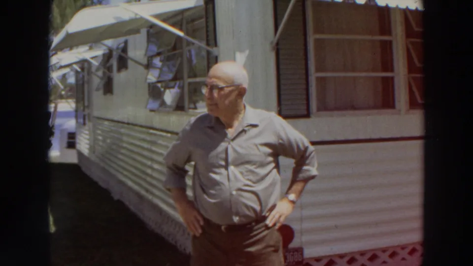 1966 Puerto Rico frame of an older man with glasses standing hands on hips beside a mobile home with open awning windows.