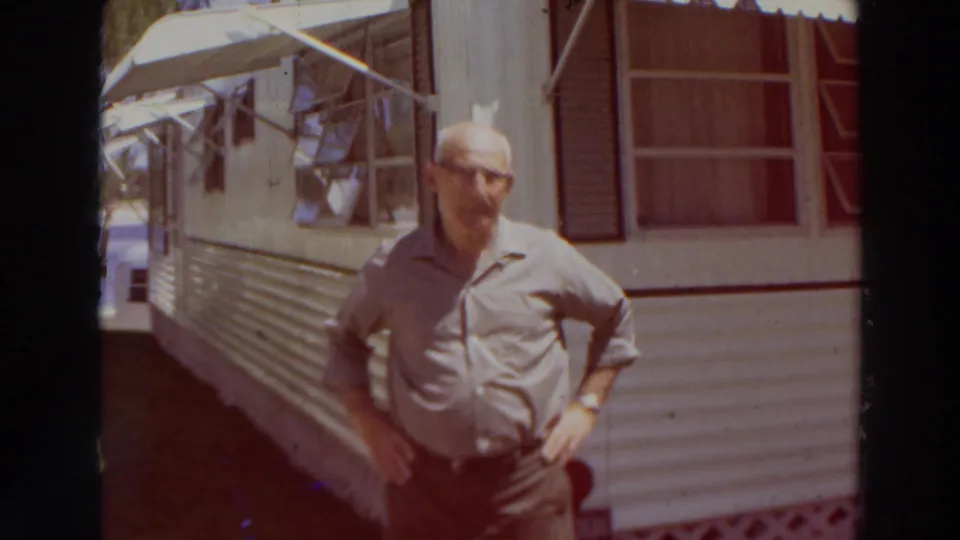 Older man remains beside the white mobile home, with awning windows, shutters, siding, and warm 8mm color shift visible.