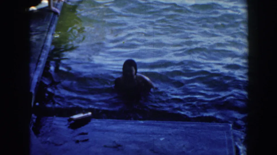 A woman continues swimming beside the dock in the Brighton, Michigan lake footage, framed by dark 8mm contact-sheet borders.