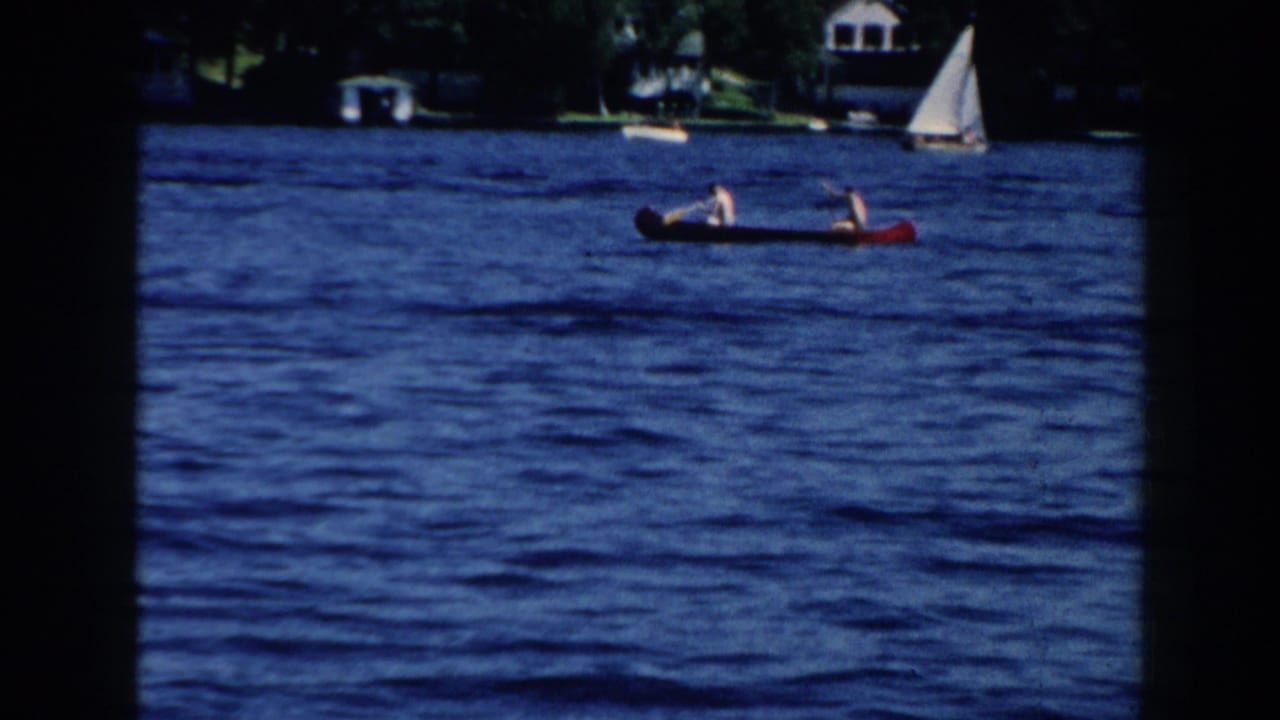 1960 Brighton, Michigan home-movie frame showing two distant people paddling a red canoe across blue lake water near a sailboat and wooded shore.