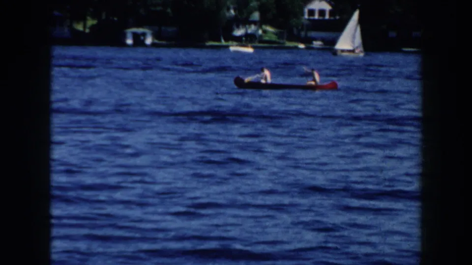 1960 Brighton, Michigan home-movie frame showing two distant people paddling a red canoe across blue lake water near a sailboat and wooded shore.