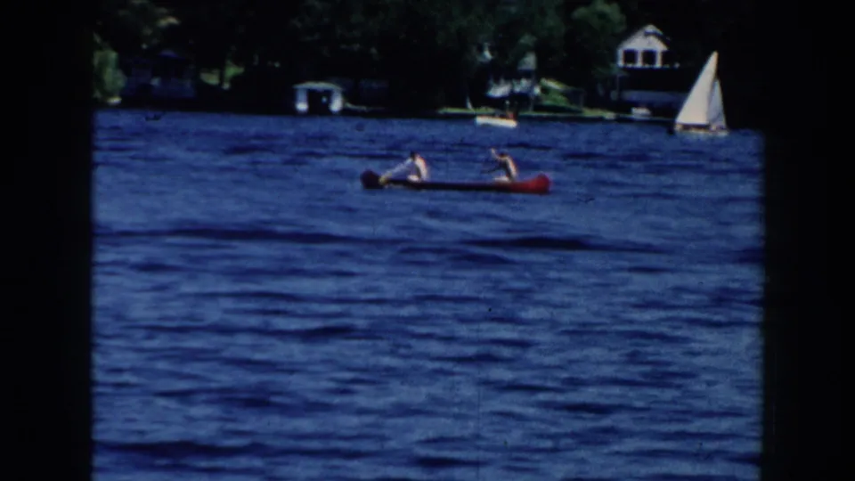 1960 Brighton, Michigan frame with a red canoe carrying two people across rippled blue lake water, with trees, houses, and a sailboat in the distance.